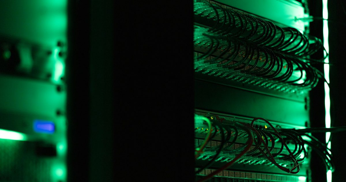 Network equipment racks lit in green in a dark room, with rows of patch panels and neatly looped network cables.
