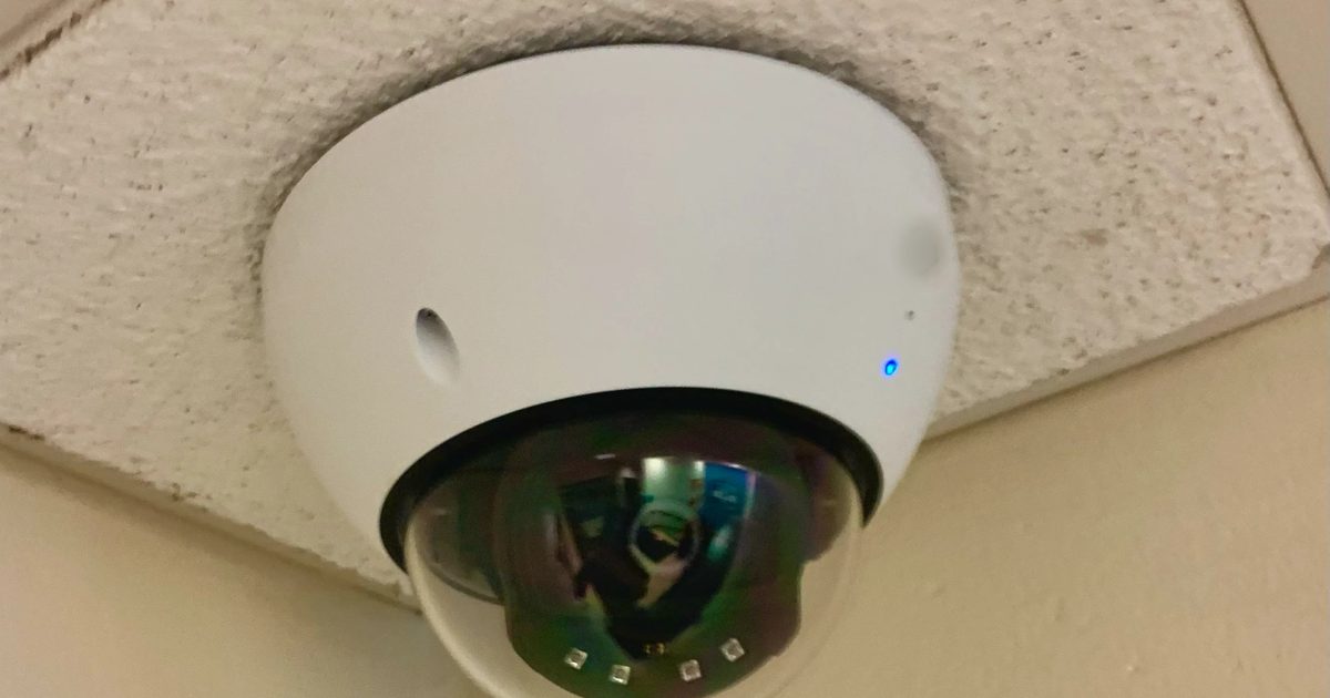 White dome security camera mounted on a textured office ceiling tile, with a small blue status light and a clear lens dome