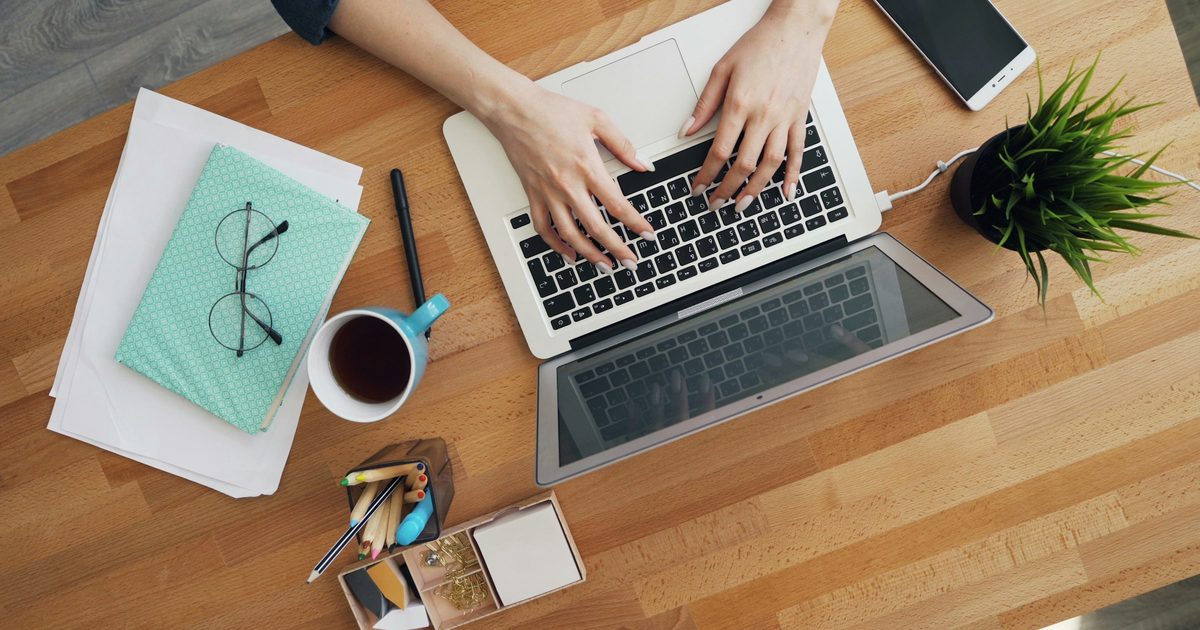 Overhead view of hands typing on a laptop on a wood desk next to a smartphone, a cup of tea, glasses on a notebook, and a small plant.
