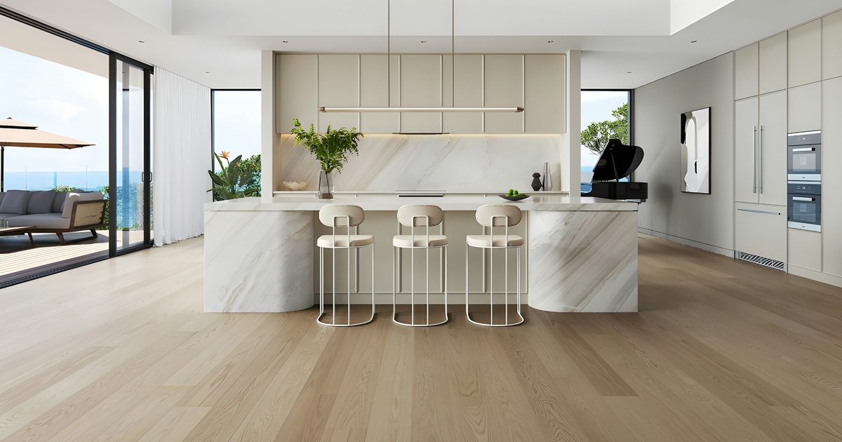 A bright open kitchen with a marble island, three cream bar stools, light oak floors and sliding glass doors to a deck with an ocean view.