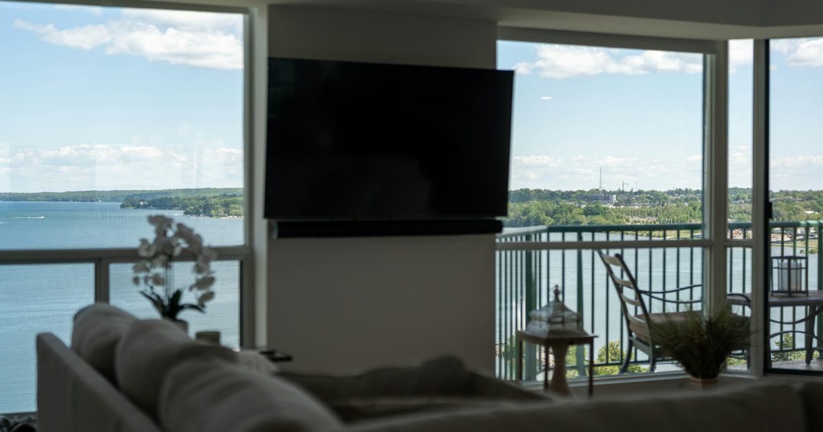 Living room with large corner windows overlooking blue water, a wall-mounted TV, a white sofa, and balcony chairs on a sunny day.