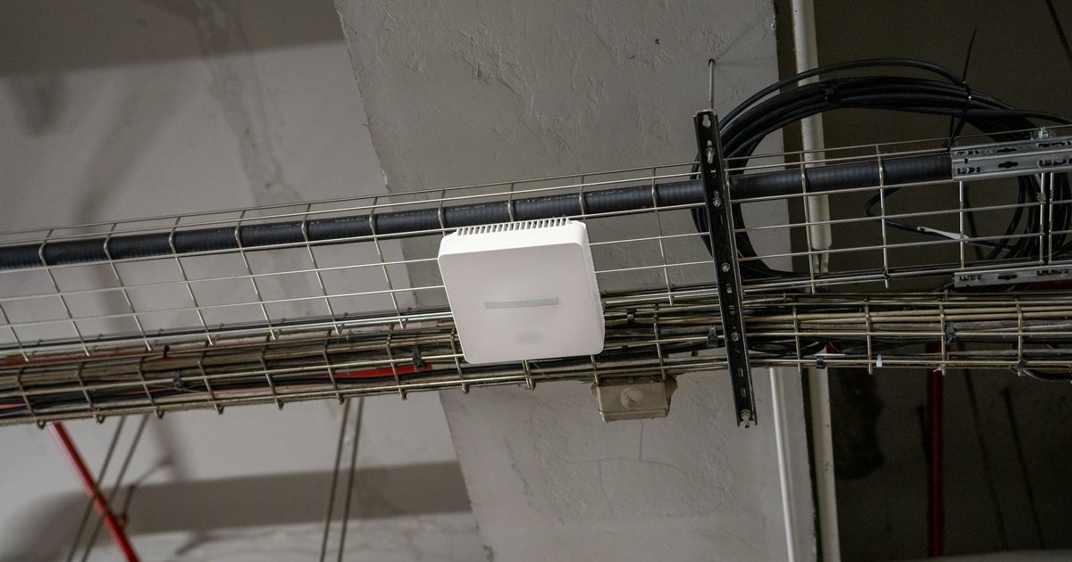 A white wireless access point mounted on a wire cable tray below a concrete ceiling, with bundled network cables running alongside.